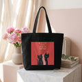 Black tote bag with cat design and text on a surface with flowers and books in the background