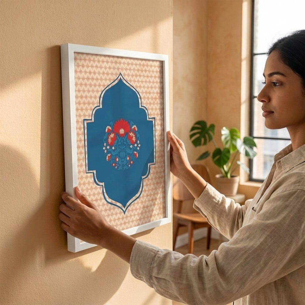 A woman in a beige linen shirt hangs a framed art print on a warm beige wall in a sunlit room. The print features a blue arched central motif with red and blue floral details, set against a beige patterned background with a white frame.