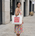 A woman in a colorful floral dress stands on a city sidewalk holding a white canvas tote bag. The bag has a pink heart graphic with the text 'EVERYTHING WORKS OUT FOR ME' in yellow and pink.