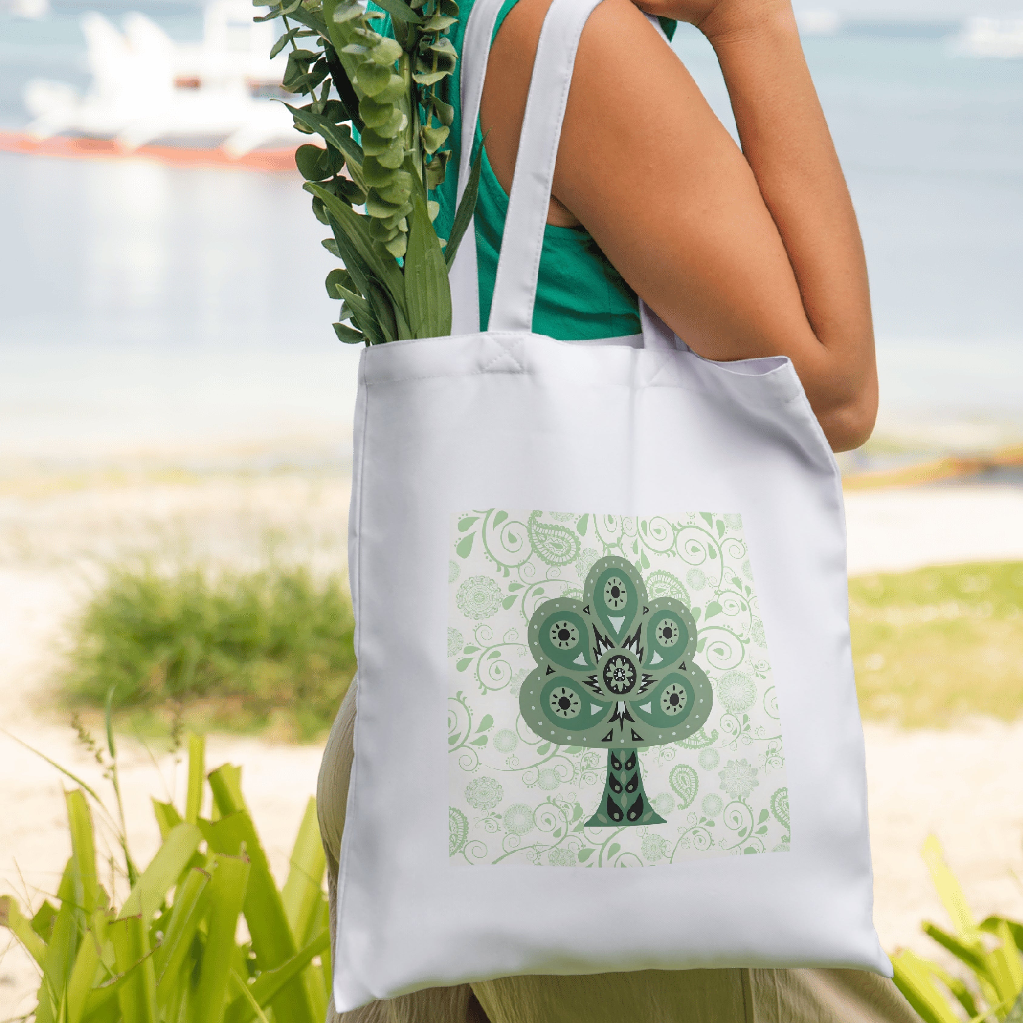 Person holding a white tote bag with a green tree design outdoors.