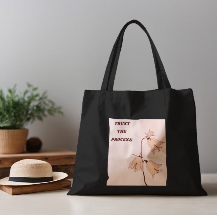 Black tote bag with floral design and 'Trust the Process' text on a neutral background