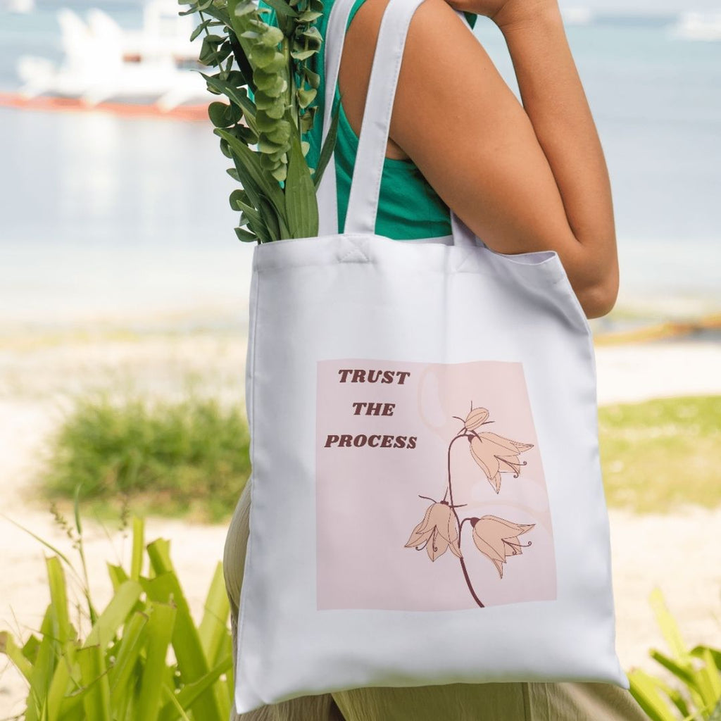 Person holding a white tote bag with a motivational quote and floral design outdoors.